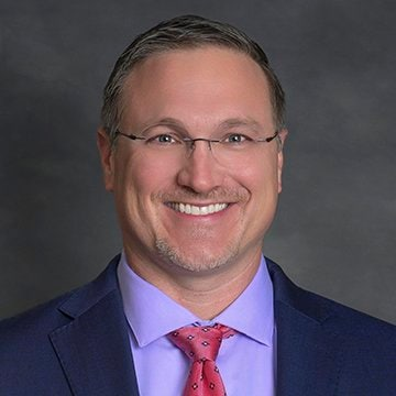 Image of a man with glasses wearing a blue shirt, red tie and blue suit jacket