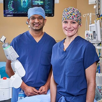 Dr. Hemil Maniar and Dr. Kelli Crabtree-Wilson stand in the operating room with the Mazor robot